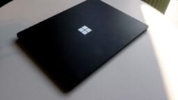 A closed black Microsoft Surface laptop sits on a round white table, partially lit by natural light.