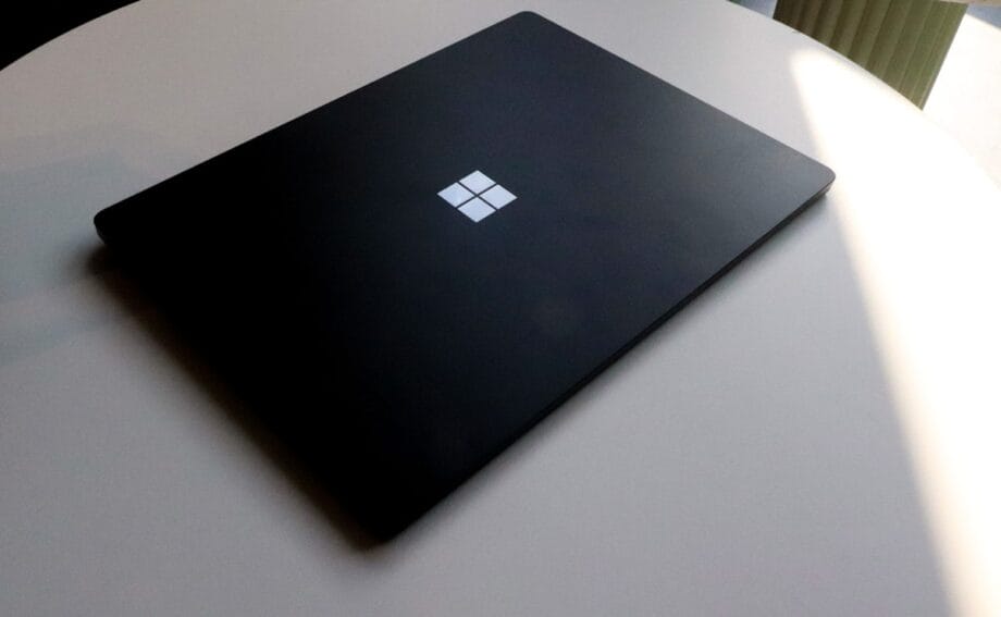 A closed black Microsoft Surface laptop sits on a round white table, partially lit by natural light.