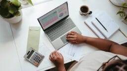 Person working at a desk with a laptop displaying charts, calculator, documents, notepad, pencil, and a cup of coffee.
