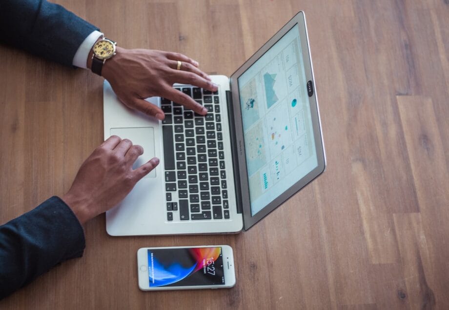 Person in a suit using a laptop with charts on the screen; a smartphone lies on the wooden table beside their hands.