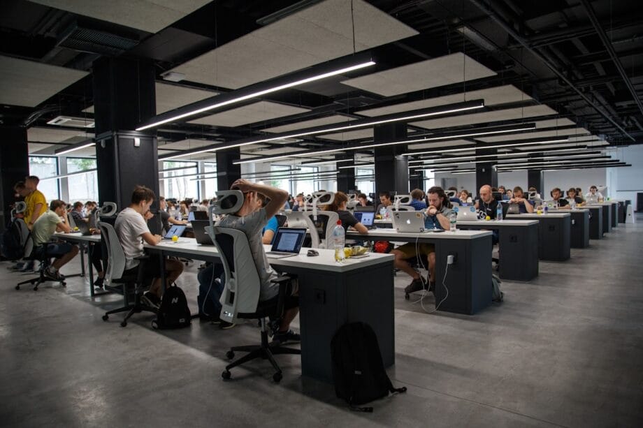 Large open-plan office with many people working at computers, seated at modern desks under bright ceiling lights.