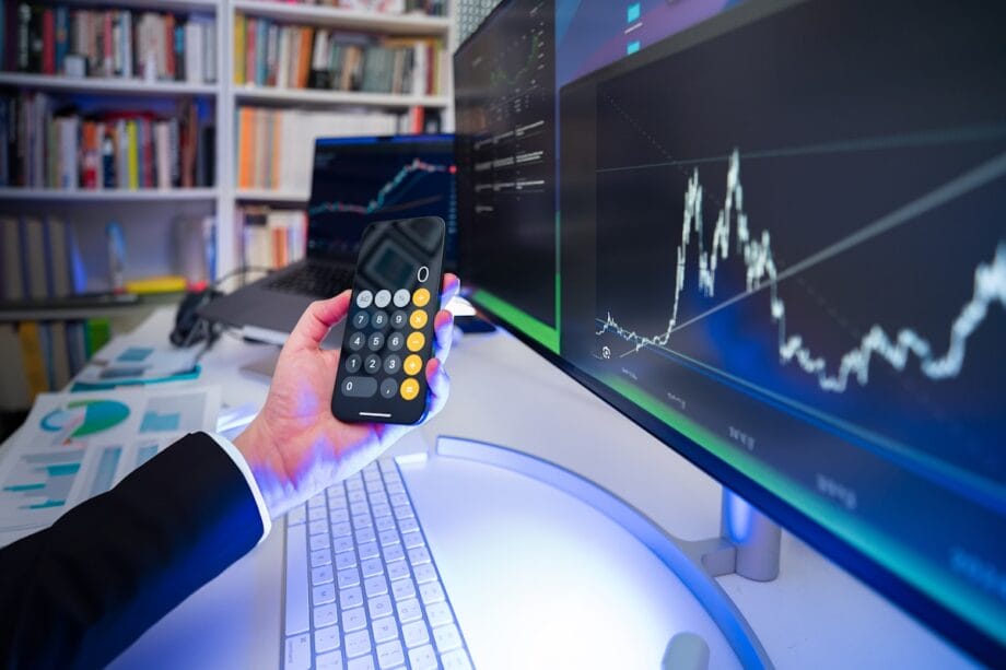 A person holds a calculator in front of computer screens displaying financial charts and graphs in an office setting.