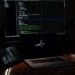 A dimly lit desk with a laptop, a tablet, a book, and a large monitor displaying code on a dark background.