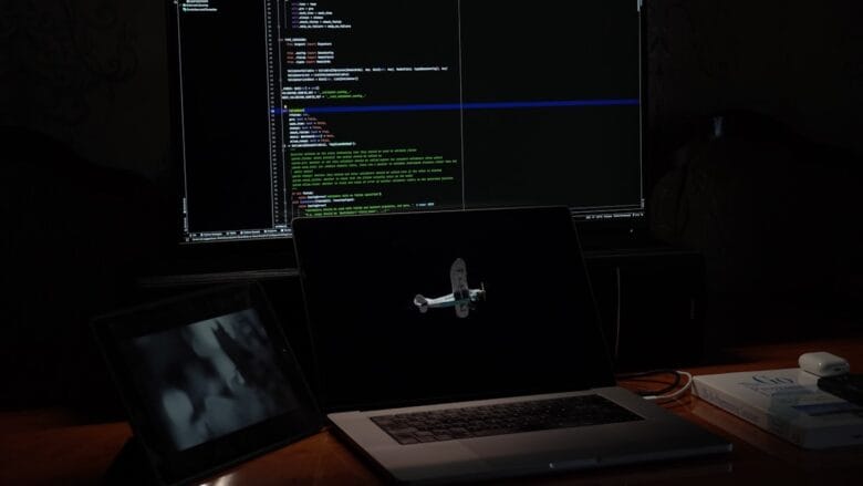 A dimly lit desk with a laptop, a tablet, a book, and a large monitor displaying code on a dark background.