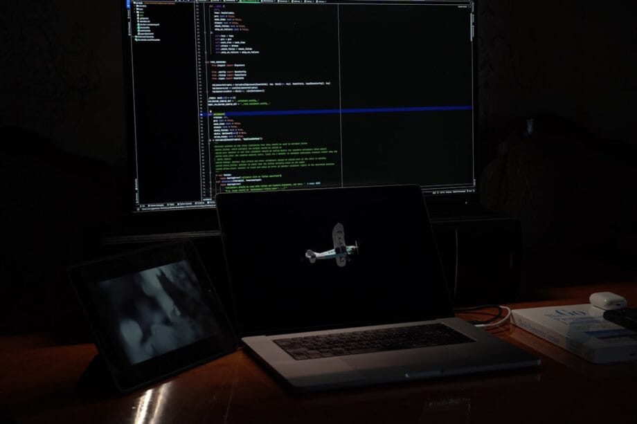 A desk with a laptop displaying an airplane model, a tablet, and a large monitor showing code in a dark room.