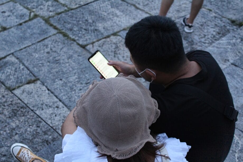 Two people sit on stone pavement, looking at a smartphone screen; other people’s legs are visible in the background.