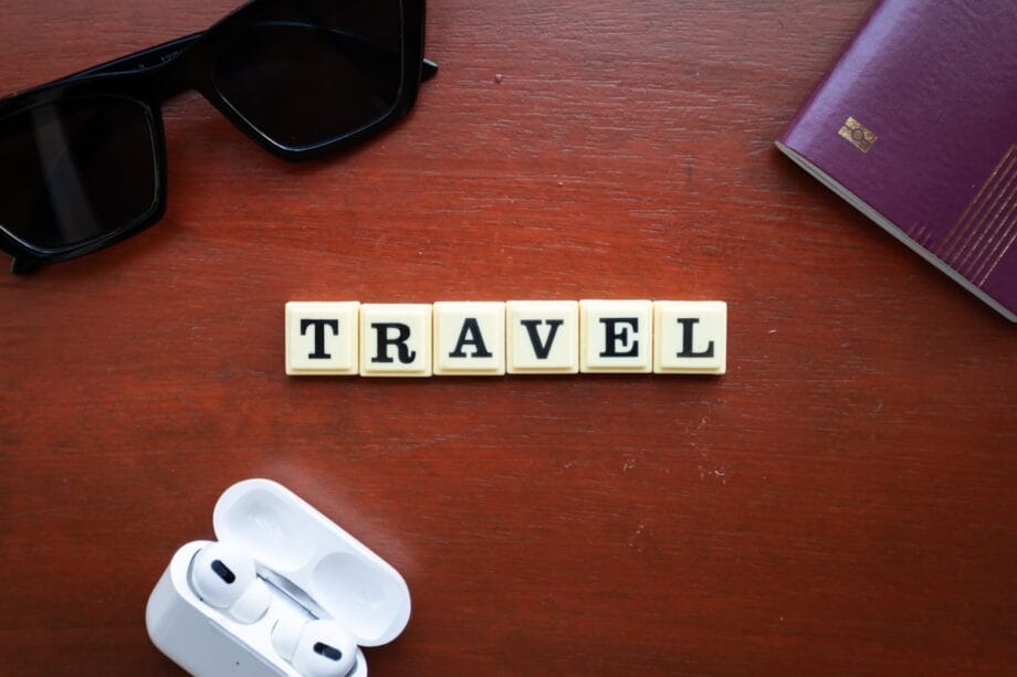 A wooden table with letter tiles spelling TRAVEL, sunglasses, wireless earbuds, and a maroon passport in the corners.