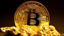 A physical Bitcoin token stands upright behind several small gold nuggets on a dark background.
