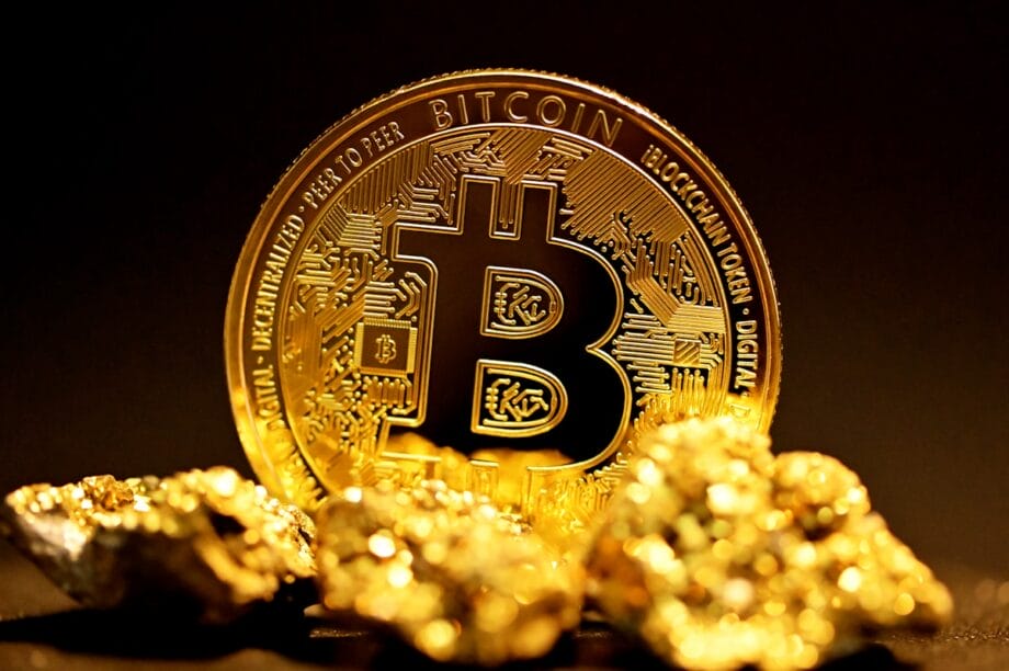 A physical Bitcoin token stands upright behind several small gold nuggets on a dark background.