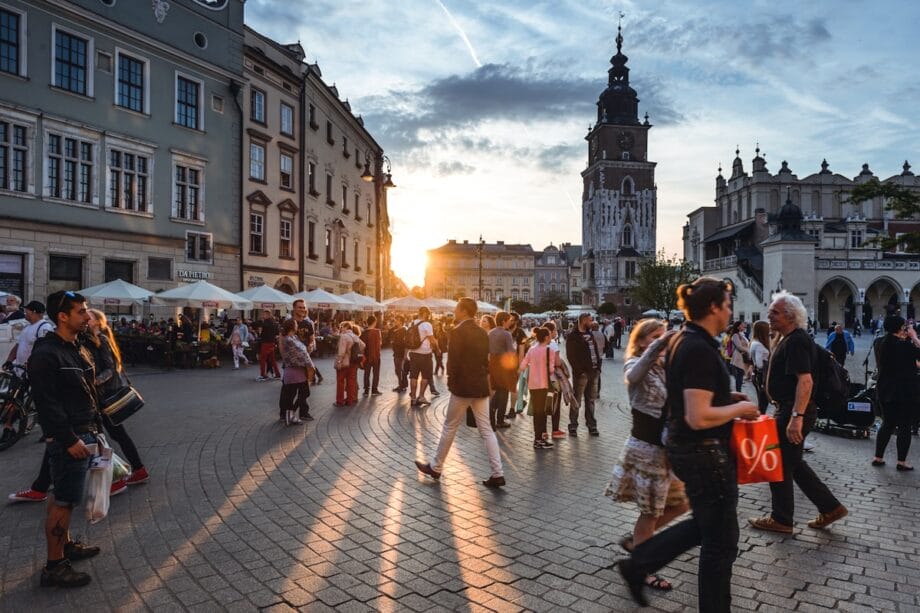 Crowd of people walking in a city square at sunset, with historic buildings and a clock tower in the background.
