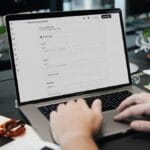 Person filling out an online form on a laptop at a desk with papers, glasses, and potted plants.