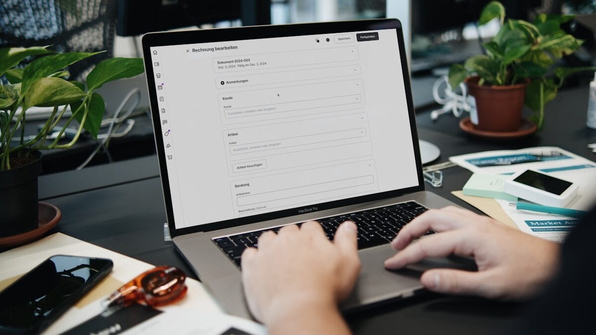 Person filling out an online form on a laptop at a desk with papers, glasses, and potted plants.