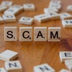 Wooden Scrabble tiles spell out SCAM on a table, surrounded by other scattered letter tiles.
