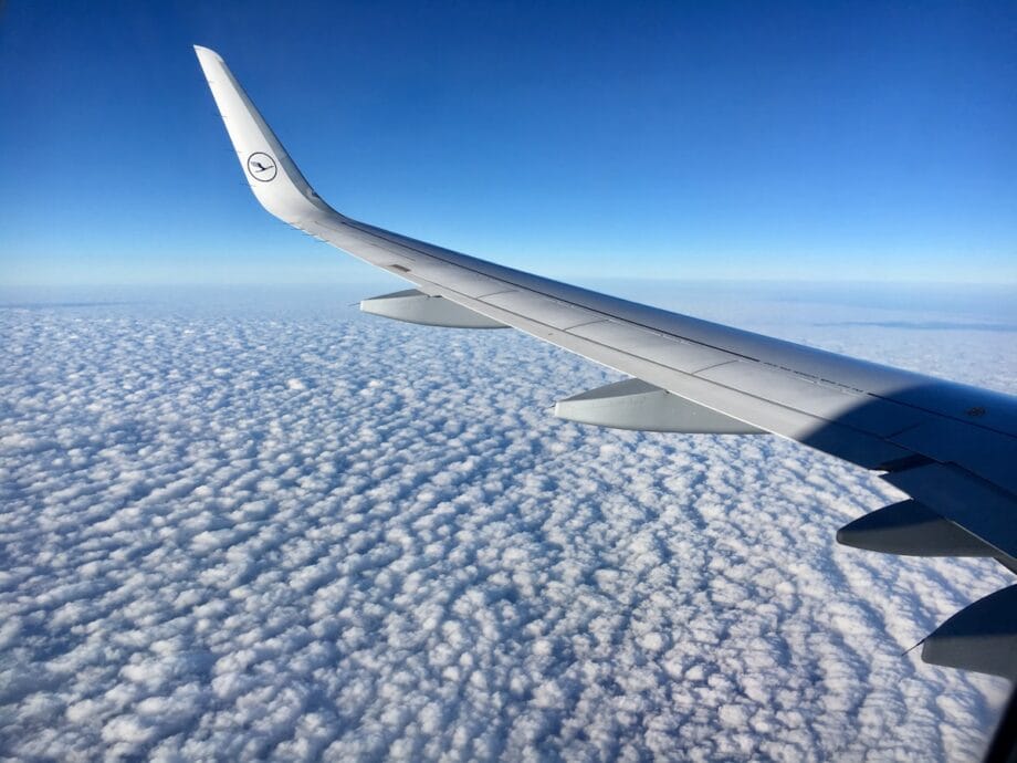 A view of an airplane wing above a blanket of clouds under a clear blue sky, taken from a passenger window.