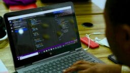 Person typing on a laptop displaying code in a text editor, with a wooden desk, cables, and other items in the background.