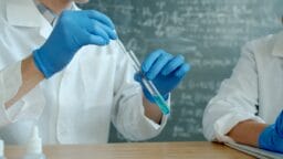 Two people in lab coats and blue gloves handling a test tube with a pipette in a laboratory, with a chalkboard in the background.