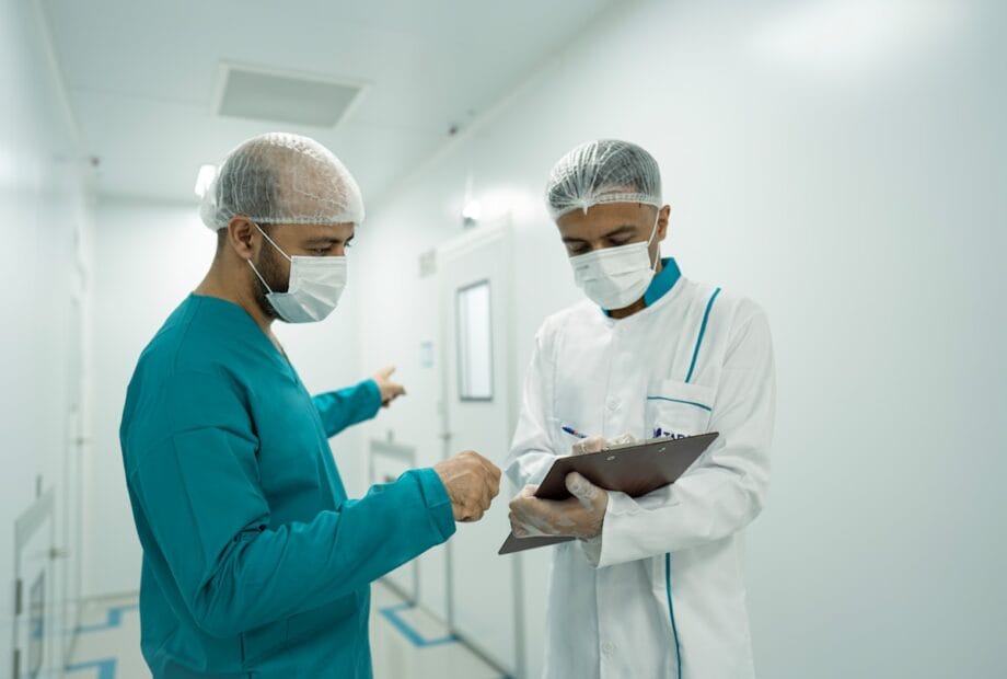 Two healthcare workers in scrubs, masks, and hairnets discuss while reviewing a clipboard in a hospital corridor.