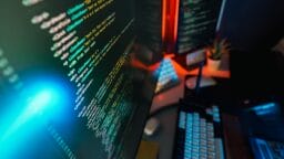 Computer screen displaying colorful lines of code, with a keyboard, mouse, and a small plant visible on the desk.