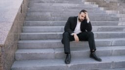 A man in a suit sits on outdoor steps, resting his head on one hand and appearing deep in thought or stressed.