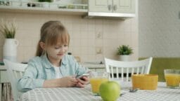 A young girl sits at a kitchen table using a smartphone, with juice, an apple, and a bowl in front of her.