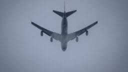 A large passenger airplane flies overhead through a cloudy, gray sky, partially obscured by mist.