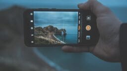 A hand holds a smartphone, taking a photo of a rocky coastline with the sea under an overcast sky.