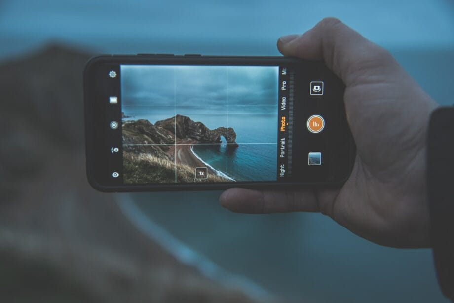 A hand holds a smartphone, taking a photo of a rocky coastline with the sea under an overcast sky.