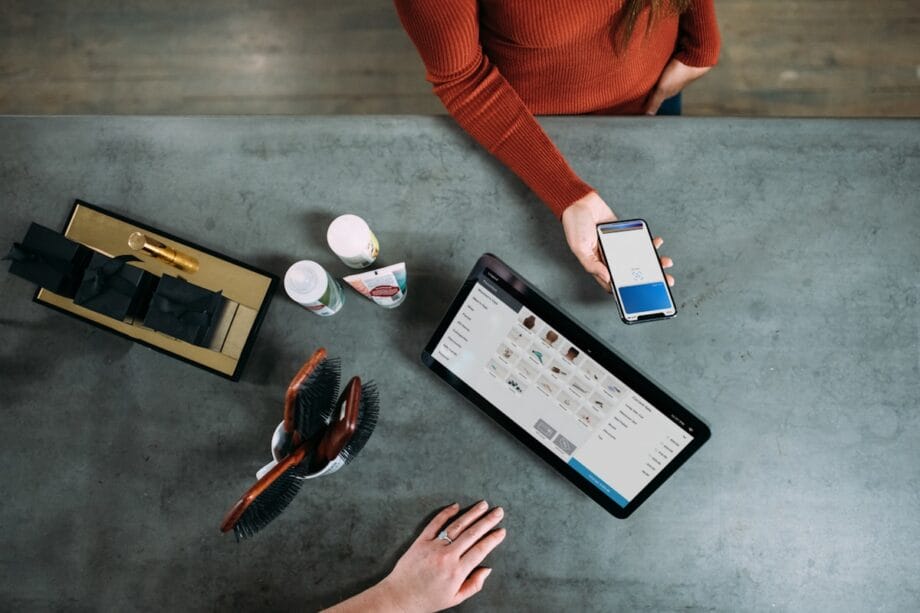 Two people at a counter; one uses a phone for payment while the other stands near a tablet displaying a product menu.