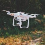 A white quadcopter drone with a camera hovers outside above grass and plants, with trees in the background.