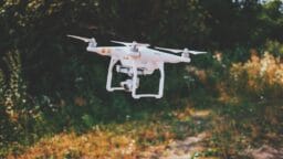 A white quadcopter drone with a camera hovers outside above grass and plants, with trees in the background.