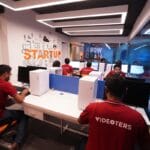 Several people wearing red “VIDEOTERS” shirts work at computers in a modern office labeled “STARTUP” with glass walls and bright lighting.