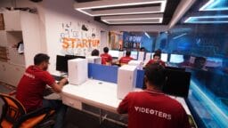 Several people wearing red “VIDEOTERS” shirts work at computers in a modern office labeled “STARTUP” with glass walls and bright lighting.