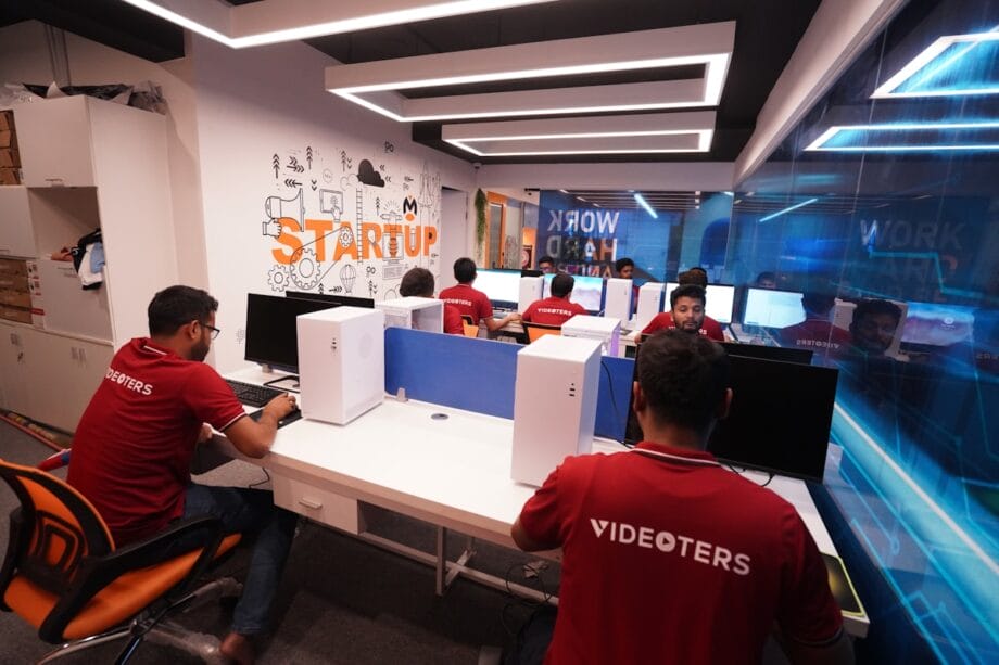 Several people wearing red “VIDEOTERS” shirts work at computers in a modern office labeled “STARTUP” with glass walls and bright lighting.