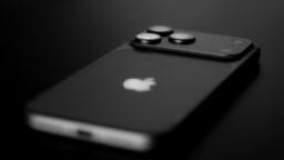 A close-up, black and white photo of a modern Apple iPhone lying face down on a dark surface.
