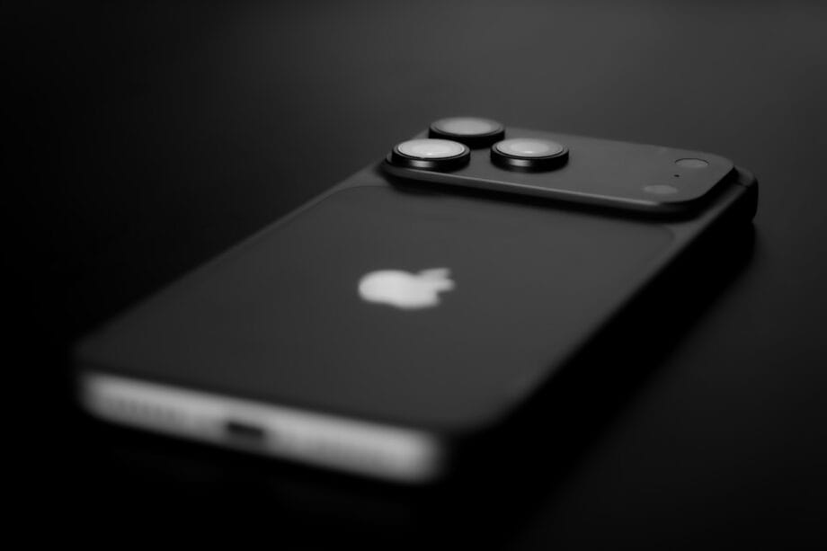 A close-up, black and white photo of a modern Apple iPhone lying face down on a dark surface.