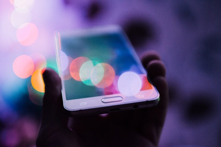 A person holds a smartphone with colorful bokeh lights reflected on its screen in a low-light setting.
