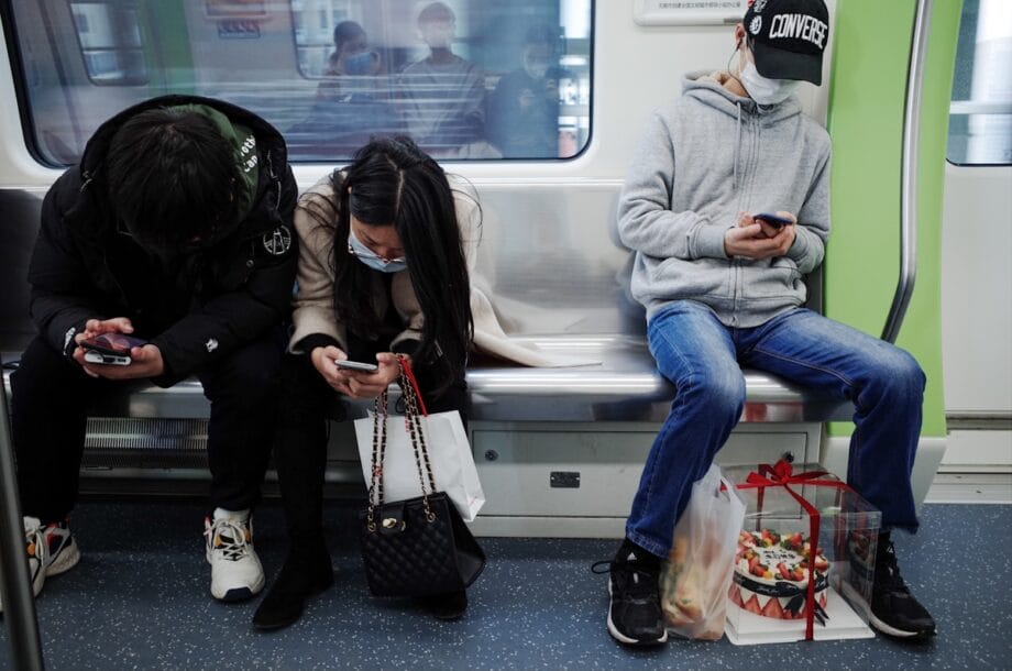 Three people wearing masks sit on a subway bench, looking at their phones; one has a gift-wrapped cake at their feet.