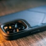 A smartphone in a black textured case rests on a wooden surface, showing its rear camera lenses closely.