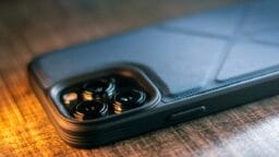 A smartphone in a black textured case rests on a wooden surface, showing its rear camera lenses closely.
