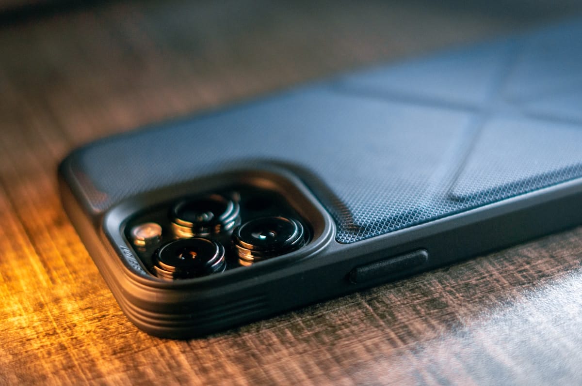 A smartphone in a black textured case rests on a wooden surface, showing its rear camera lenses closely.