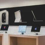 Three Apple iMac computers are displayed on a counter beneath a large poster showing an iPad and accessories in a retail store.