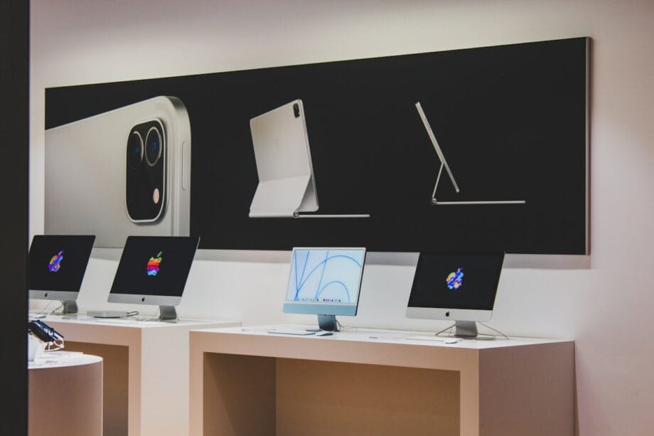 Three Apple iMac computers are displayed on a counter beneath a large poster showing an iPad and accessories in a retail store.