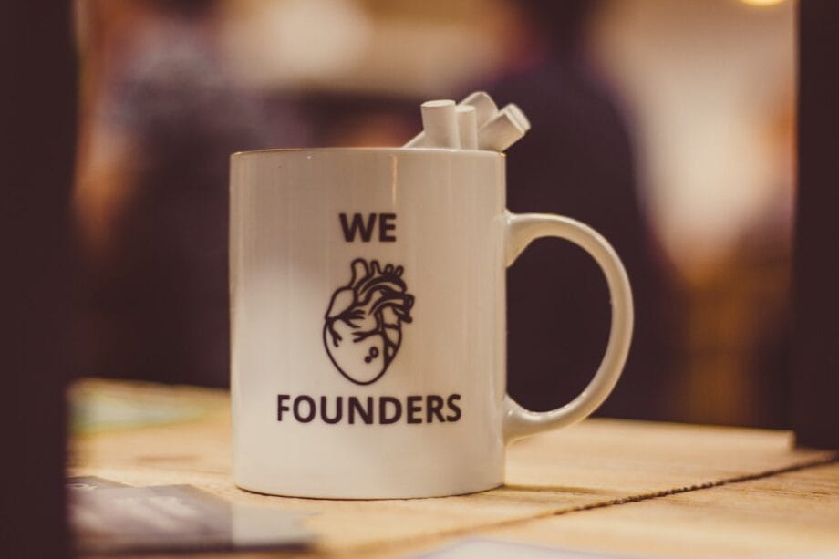White mug with an anatomical heart illustration and the words WE FOUNDERS, holding three white straws, placed on a wooden surface.