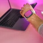 A person wearing a smartwatch uses a smartphone in front of a laptop on a pink desk with a wireless mouse and earbuds nearby.