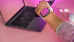 A person wearing a smartwatch uses a smartphone in front of a laptop on a pink desk with a wireless mouse and earbuds nearby.