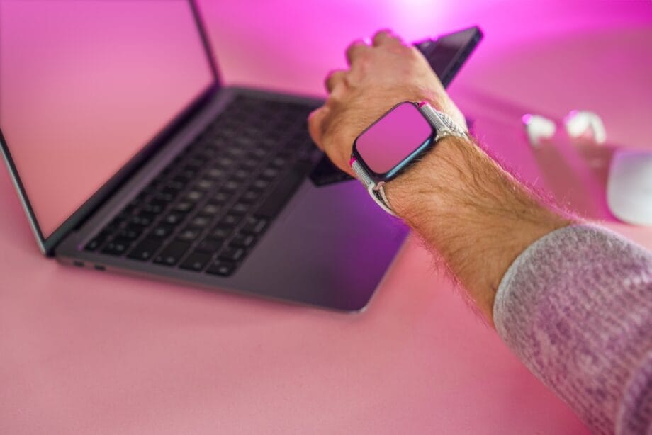 A person wearing a smartwatch uses a smartphone in front of a laptop on a pink desk with a wireless mouse and earbuds nearby.