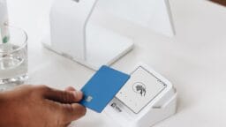 A hand holds a blue credit card near a white contactless payment terminal on a table.