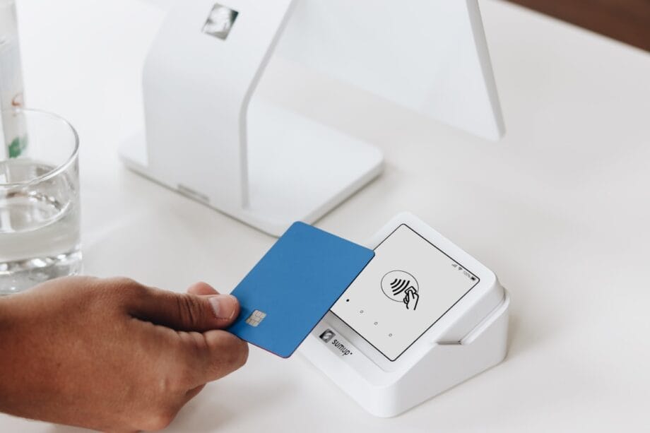 A hand holds a blue credit card near a white contactless payment terminal on a table.