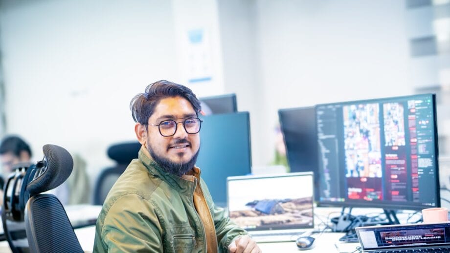 Man with glasses sitting at a desk in an office, looking at the camera, with computer monitors and a laptop in front of him.