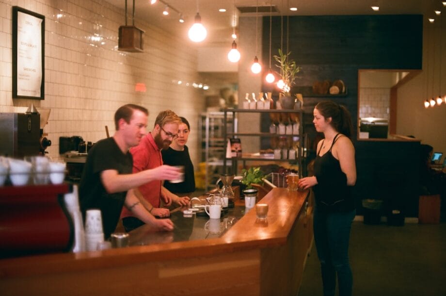 Four people stand at a caf&eacute; counter, with one person serving drinks to a woman who is waiting on the other side.